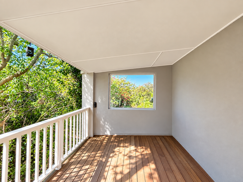 A clean, well-maintained covered balcony featuring timber decking, a white balustrade, and a fixed window overlooking lush greenery.