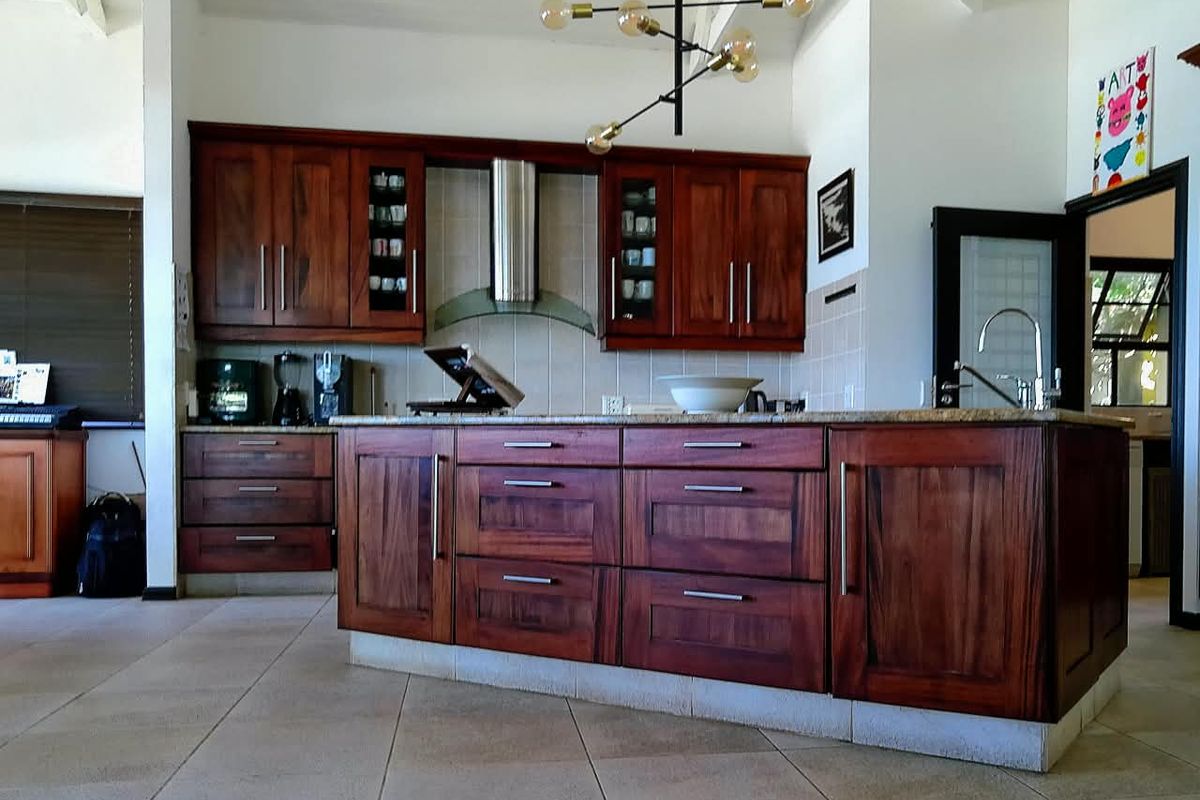 A spacious kitchen featuring rich wooden cabinetry, a large central island with granite countertops, stainless steel extractor fan, and neutral tiled flooring.