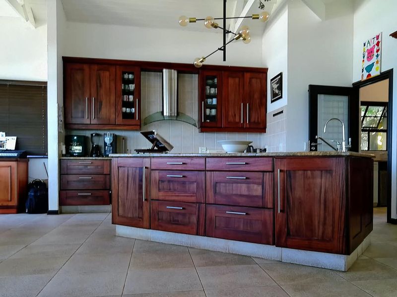 A spacious kitchen featuring rich wooden cabinetry, a large central island with granite countertops, stainless steel extractor fan, and neutral tiled flooring.