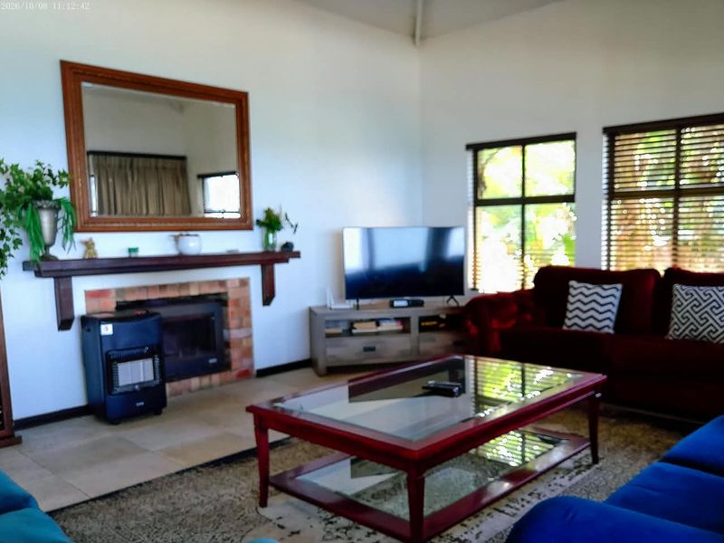 A comfortable living room featuring a brick fireplace with a wooden mantelpiece, large windows with venetian blinds, and high ceilings.