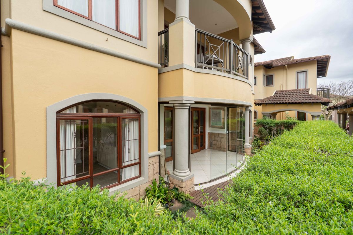 A well-maintained rear exterior view featuring a covered patio with glass enclosure, balcony, stone cladding, and a neatly hedged garden.