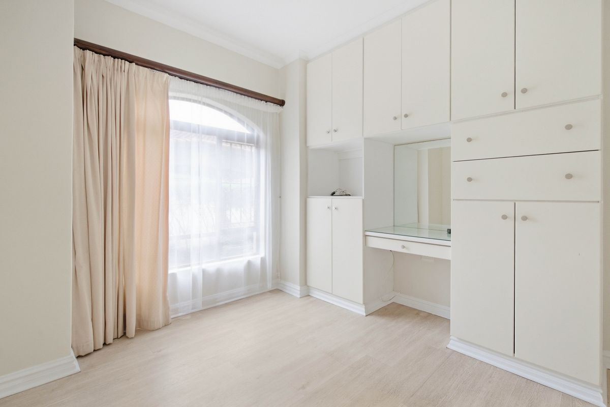 A bright, empty bedroom featuring extensive built-in cabinetry with an integrated vanity unit, arched window, and light-toned laminate flooring.