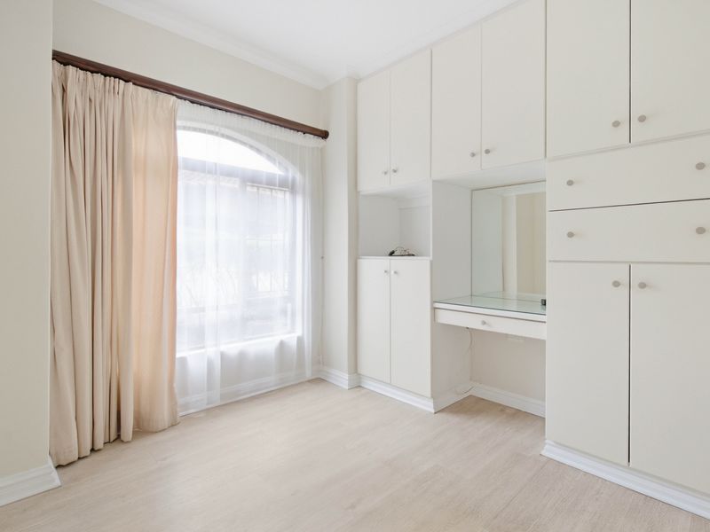 A bright, empty bedroom featuring extensive built-in cabinetry with an integrated vanity unit, arched window, and light-toned laminate flooring.