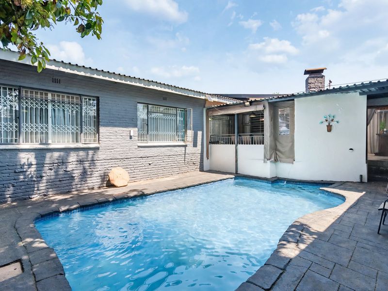 A private outdoor swimming pool area featuring a paved deck, brick exterior walls with security bars, and a chimney stack.