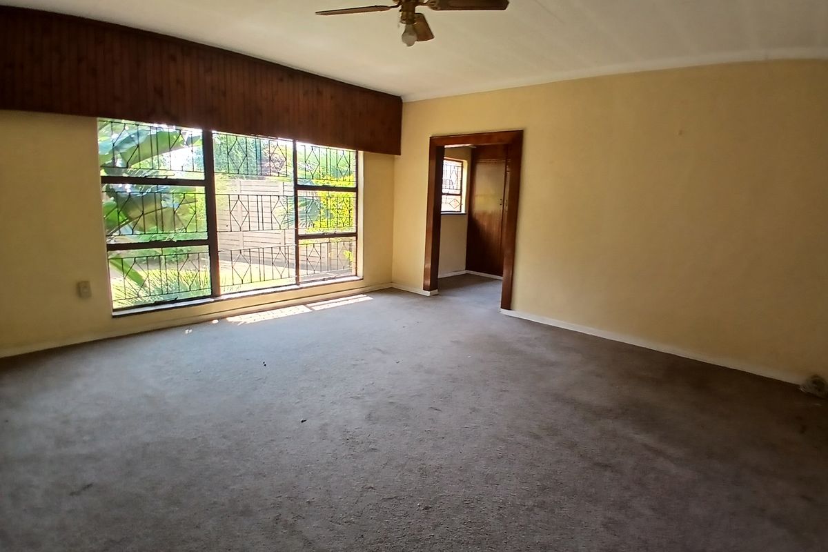 Spacious, empty living room featuring carpet flooring, a ceiling fan, and large windows with security bars, offering a blank canvas for renovation.