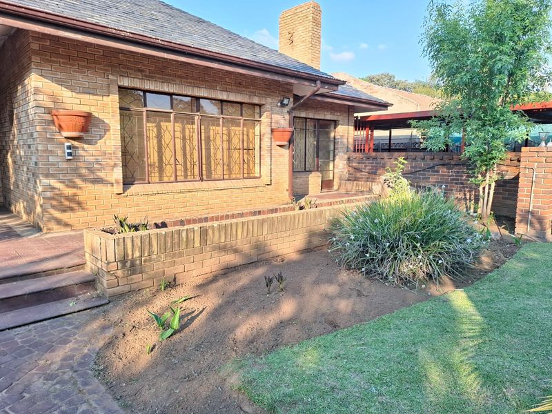 Rear view of a brick house featuring a chimney, paved walkway, and a landscaped garden area with a low brick retaining wall.