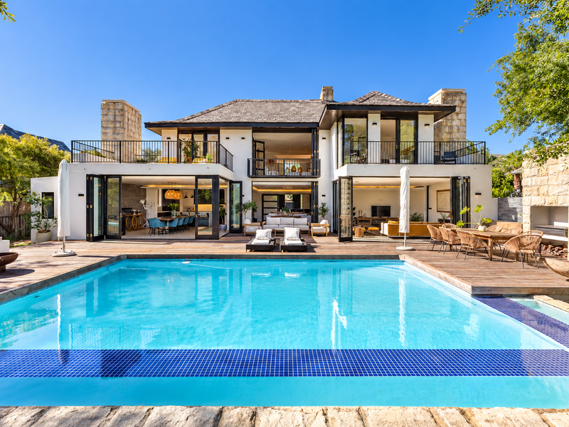 A stunning rear exterior view featuring a large swimming pool, expansive timber decking, and a modern home with bi-fold doors and stone-clad architectural details.
