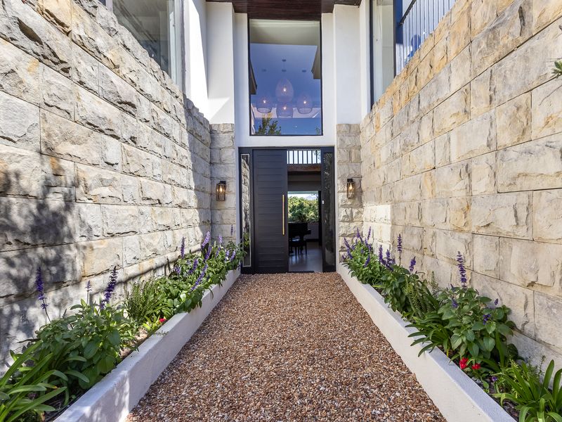 A striking entrance featuring a gravel pathway flanked by stone-clad walls and built-in planters, leading to a modern front door with a large glass window.
