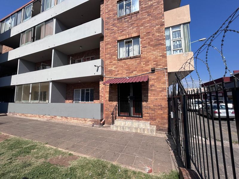 Front exterior view of a multi-story face brick townhouse complex featuring security fencing, paved walkways, and private balconies.