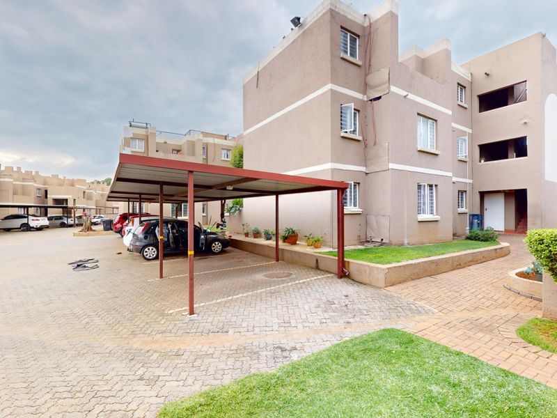 Exterior view of a multi-story townhouse complex featuring a paved driveway, covered carport parking, and established garden areas.