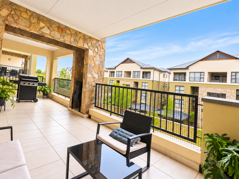 Spacious covered balcony featuring stone cladding, tiled flooring, and a metal balustrade, offering an open-air space with views of the surrounding apartment complex.