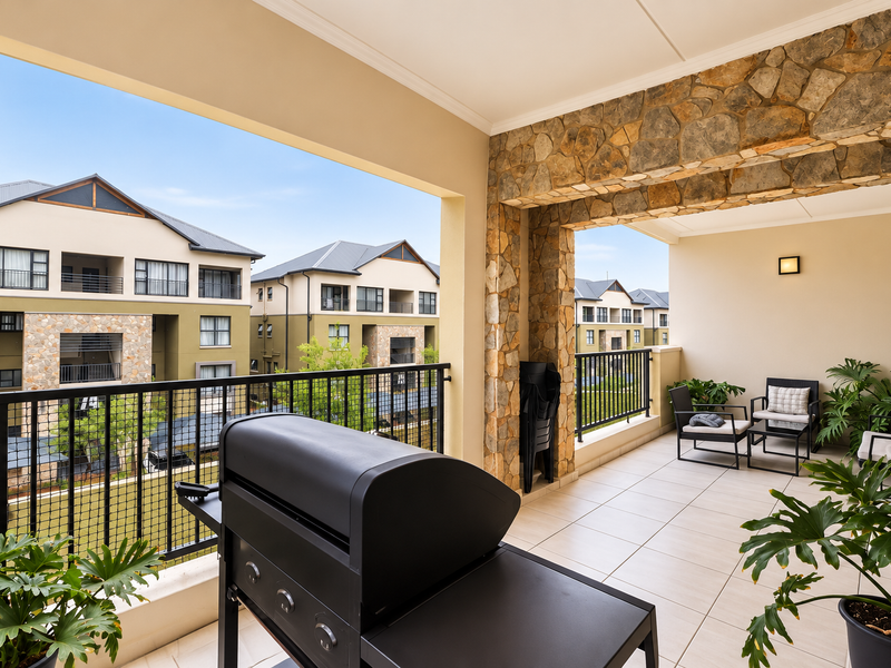 A spacious covered balcony featuring stone-clad pillars, tiled flooring, and a metal balustrade, offering an open view of the surrounding apartment complex.