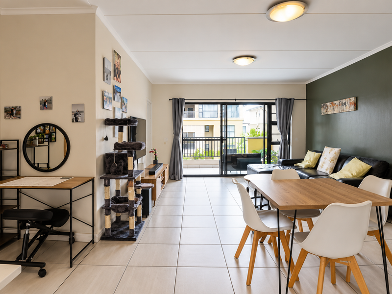 An open-plan living and dining area featuring neutral tiled flooring, an olive green accent wall, and large sliding doors leading to a private balcony.