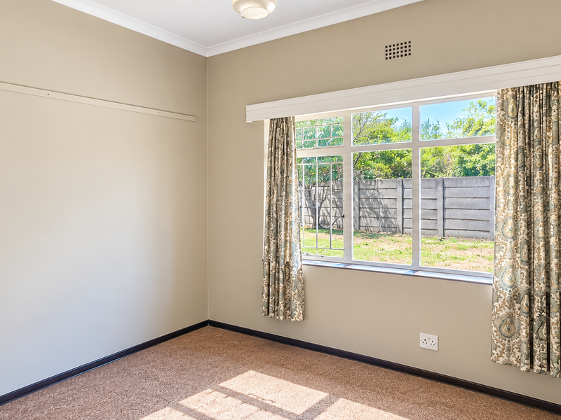 A bright, empty bedroom featuring neutral wall tones, carpeted flooring, and a large window overlooking the rear garden area.