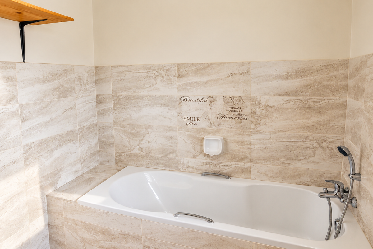 A clean bathroom featuring a built-in bathtub with chrome fixtures, surrounded by neutral-toned ceramic wall tiles and a convenient wall-mounted shelf.