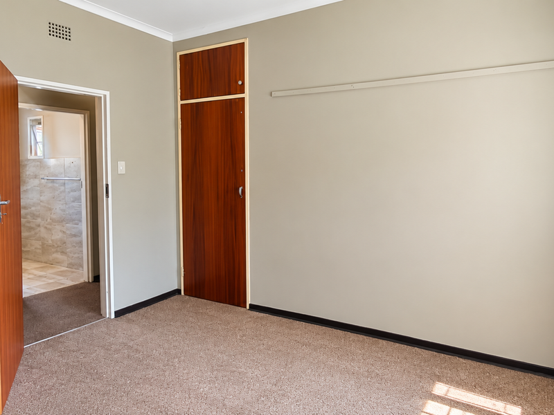A vacant bedroom featuring neutral wall tones, carpeted flooring, and a built-in wooden cupboard, with an adjacent view into a tiled bathroom.