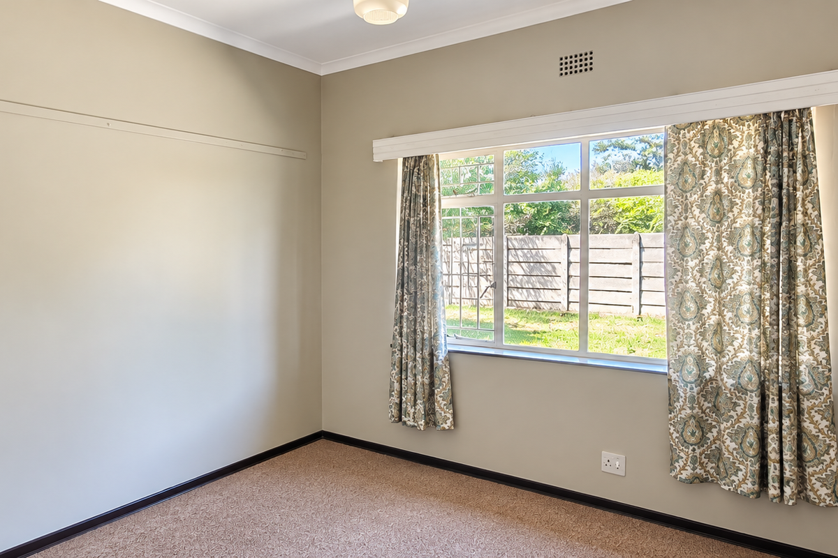 A bright, empty bedroom featuring neutral wall tones, carpeted flooring, and a large window with a curtain rail, offering a clean canvas for personalization.