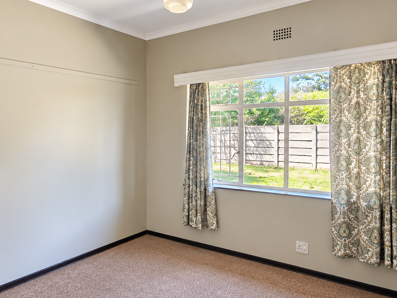 A bright, empty bedroom featuring neutral wall tones, carpeted flooring, and a large window with a curtain rail, offering a clean canvas for personalization.