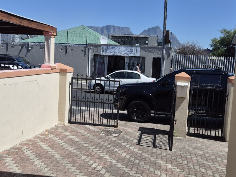 Street view showing a paved driveway entrance with a metal security gate and masonry boundary wall, with mountain views in the background.
