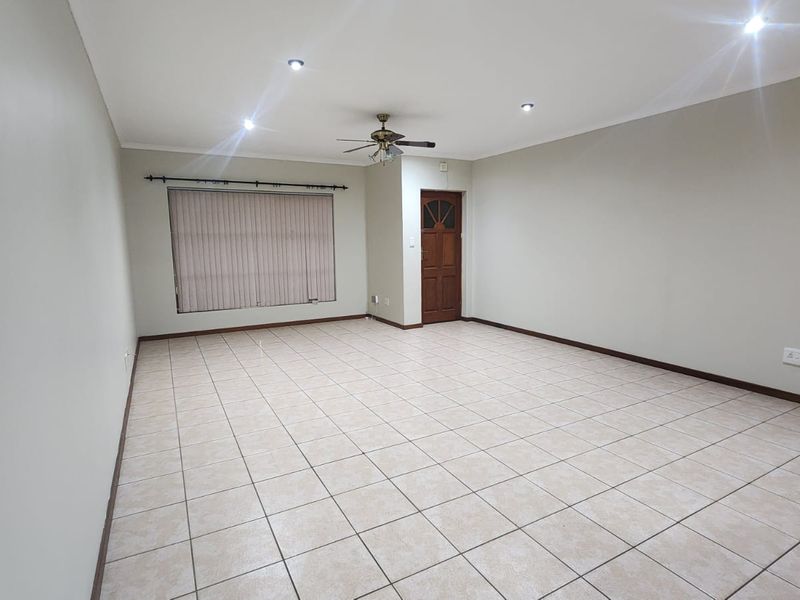 A spacious, empty living area featuring neutral tiled flooring, recessed downlighting, a ceiling fan, and a timber entrance door.