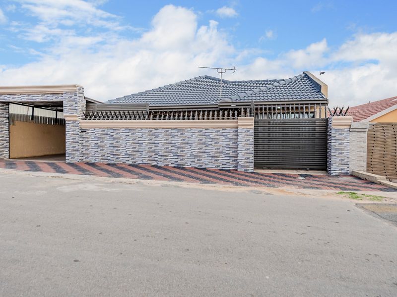 Front exterior view of the house featuring a tiled roof, secure perimeter wall with spikes, sliding gate, and a paved driveway with a carport.
