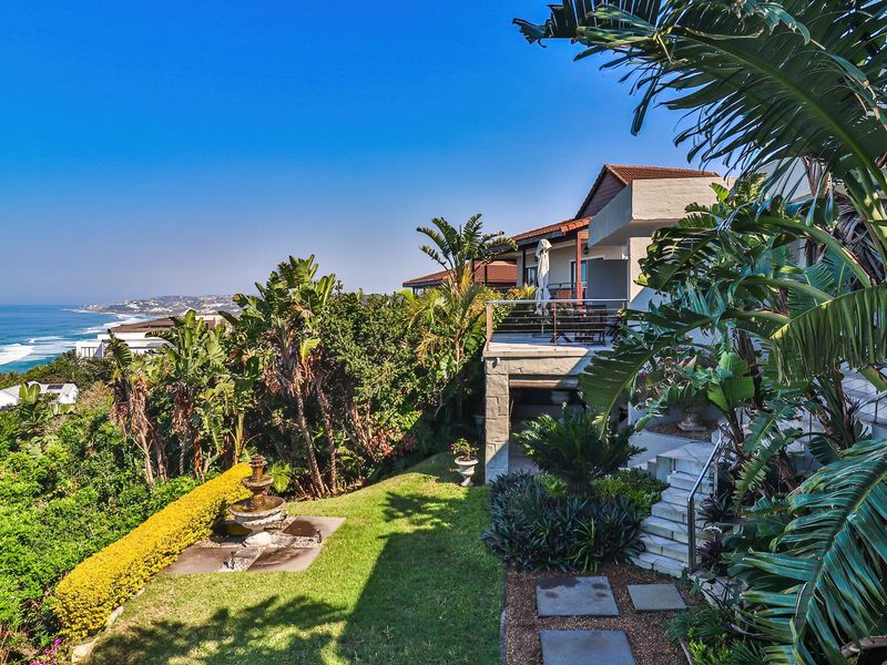 A scenic rear exterior view featuring a landscaped garden, stone fountain, and a balcony overlooking the ocean.