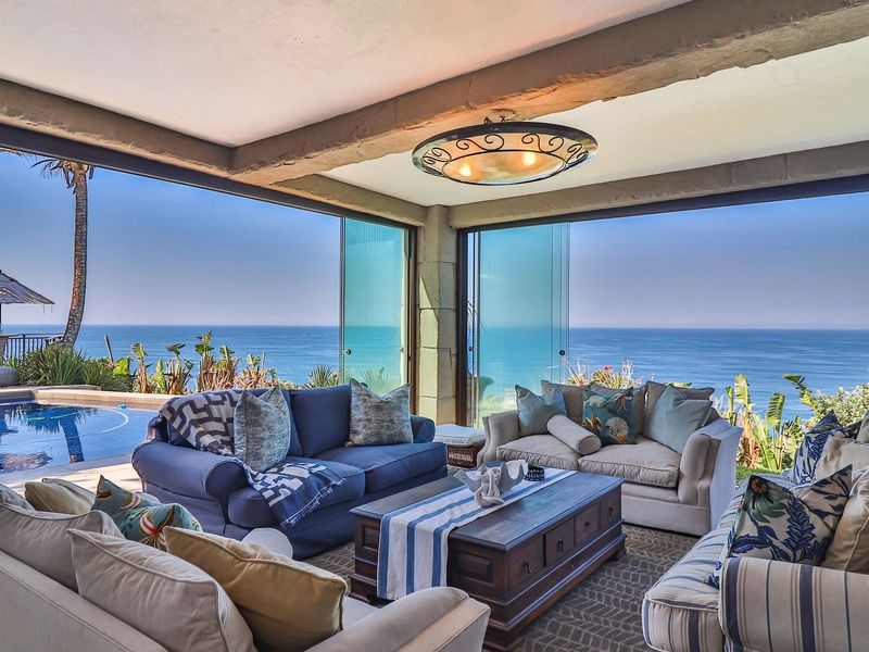 Covered patio featuring expansive stacking glass doors, exposed stone ceiling beams, and direct access to the pool area with panoramic ocean views.