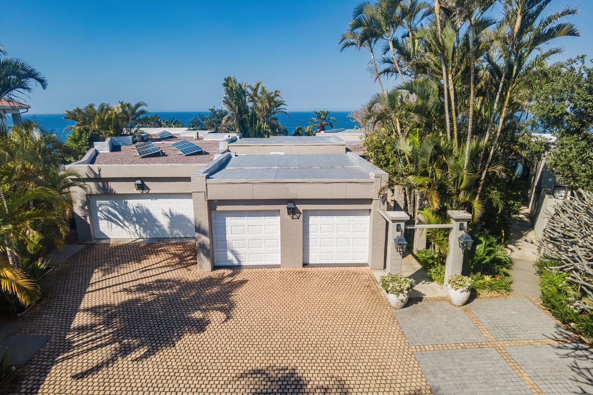 Aerial view of a coastal property featuring a triple garage, paved driveway, and roof-mounted solar panels set within a lush, tropical garden.