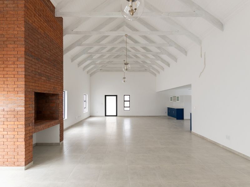 Spacious open-plan living area featuring a striking exposed brick fireplace, vaulted ceilings with timber trusses, and neutral tiled flooring throughout.