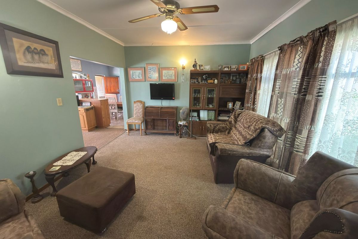 A spacious living room featuring carpeted flooring, a ceiling fan, and coved cornicing, with an open-plan flow leading into the adjacent kitchen area.