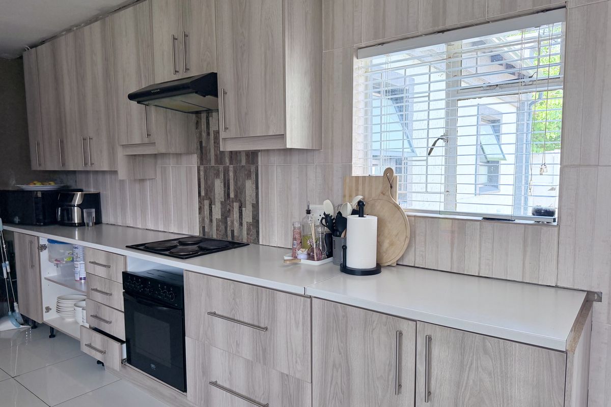 A functional kitchen featuring light wood-grain cabinetry, integrated electric hob and oven, tiled splashbacks, and tiled flooring with a window overlooking the exterior.