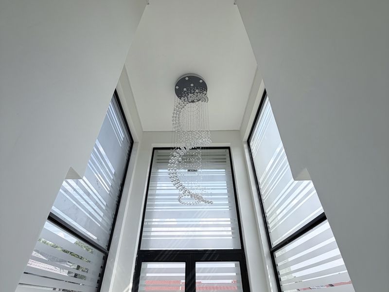 A bright, double-height entrance foyer featuring expansive glass windows, a modern glass door, and a contemporary chandelier.
