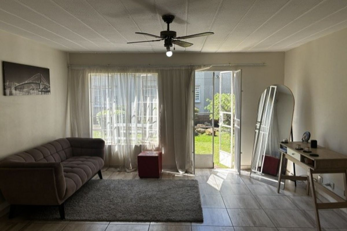 A bright living room featuring tiled flooring, a ceiling fan, and direct access to a private garden through a glass door with security gate.