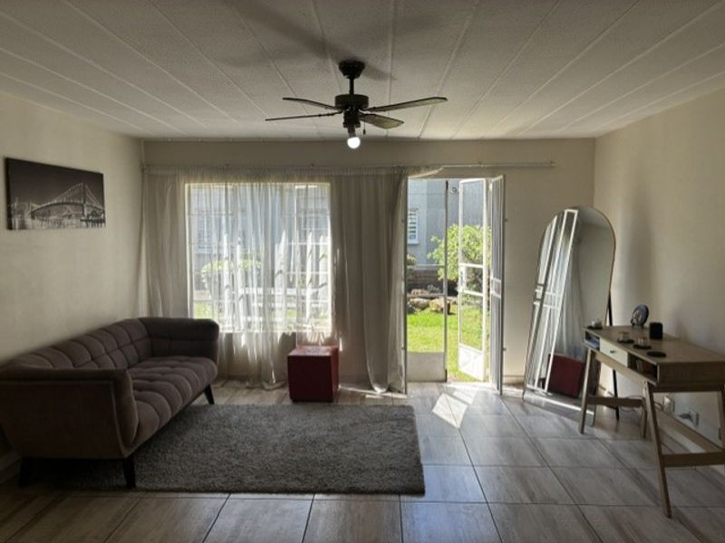 A bright living room featuring tiled flooring, a ceiling fan, and direct access to a private garden through a glass door with security gate.