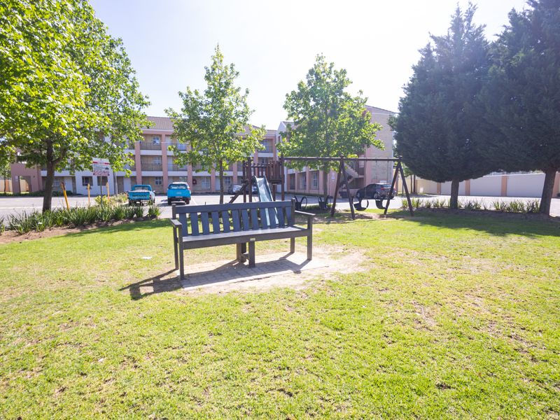 A communal garden area featuring a lawn, mature trees, a park bench, and playground equipment, situated within an apartment complex.