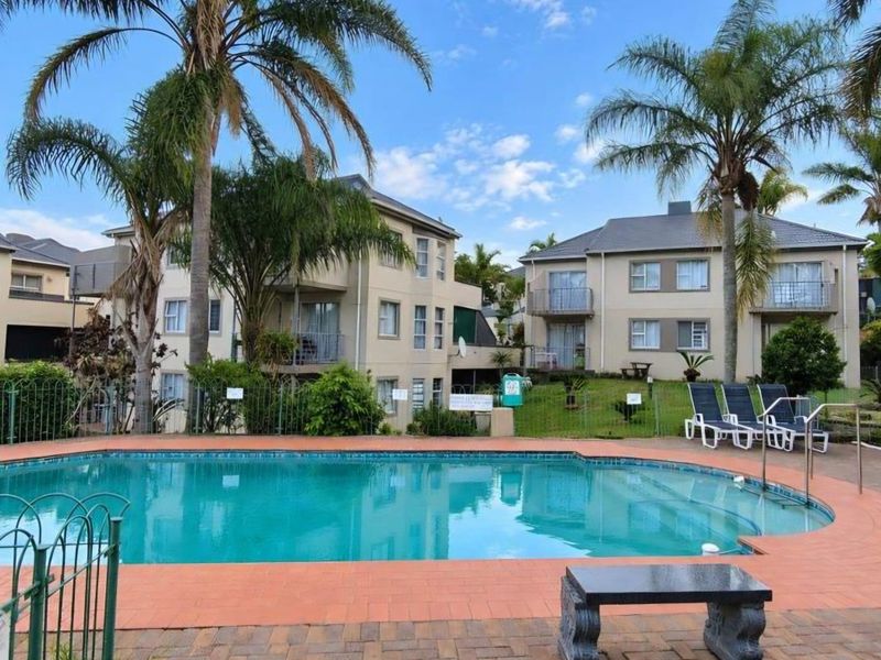 Communal swimming pool area featuring a paved deck and metal fencing, set within a well-maintained townhouse complex with multi-story residential units.