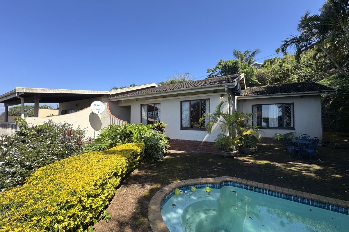 Rear exterior view of the house featuring a swimming pool, covered patio area, and established garden surroundings.