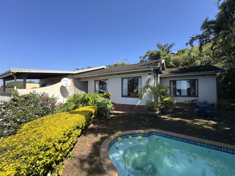 Rear exterior view of the house featuring a swimming pool, covered patio area, and established garden surroundings.