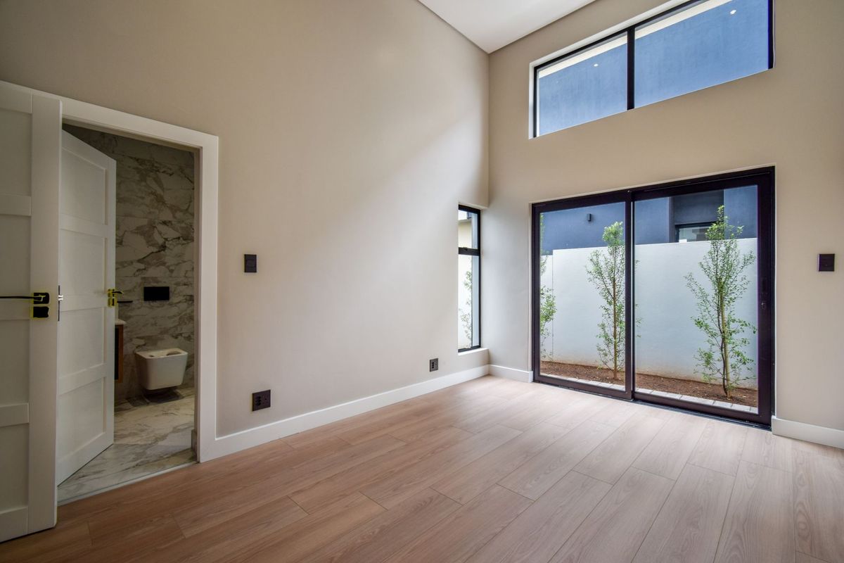 A bright, modern bedroom featuring high ceilings, light wood-look flooring, large sliding glass doors, and an en-suite bathroom with marble-style tiling.