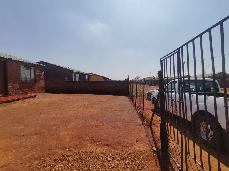 Front exterior view of a residential property featuring a brick facade, metal security gate, and an unpaved yard area enclosed by a boundary wall.