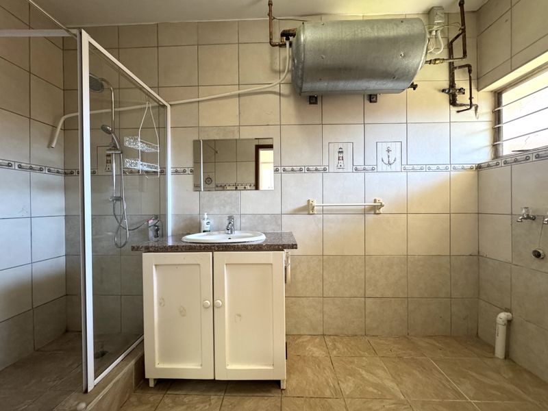 A dated bathroom featuring a shower enclosure, vanity basin, and exposed geyser unit with tiled finishes throughout.