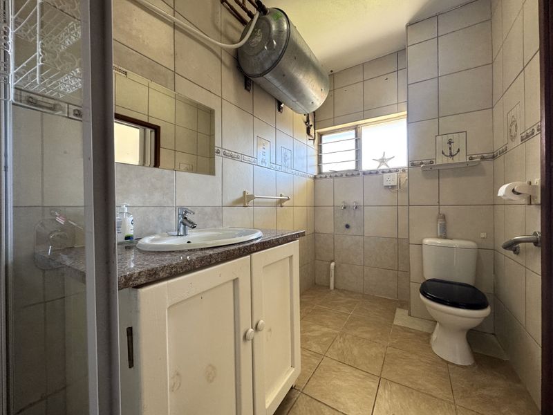 A functional bathroom featuring a vanity with basin, toilet, and exposed geyser. The space is fully tiled and requires cosmetic updates to modernize the finishes.