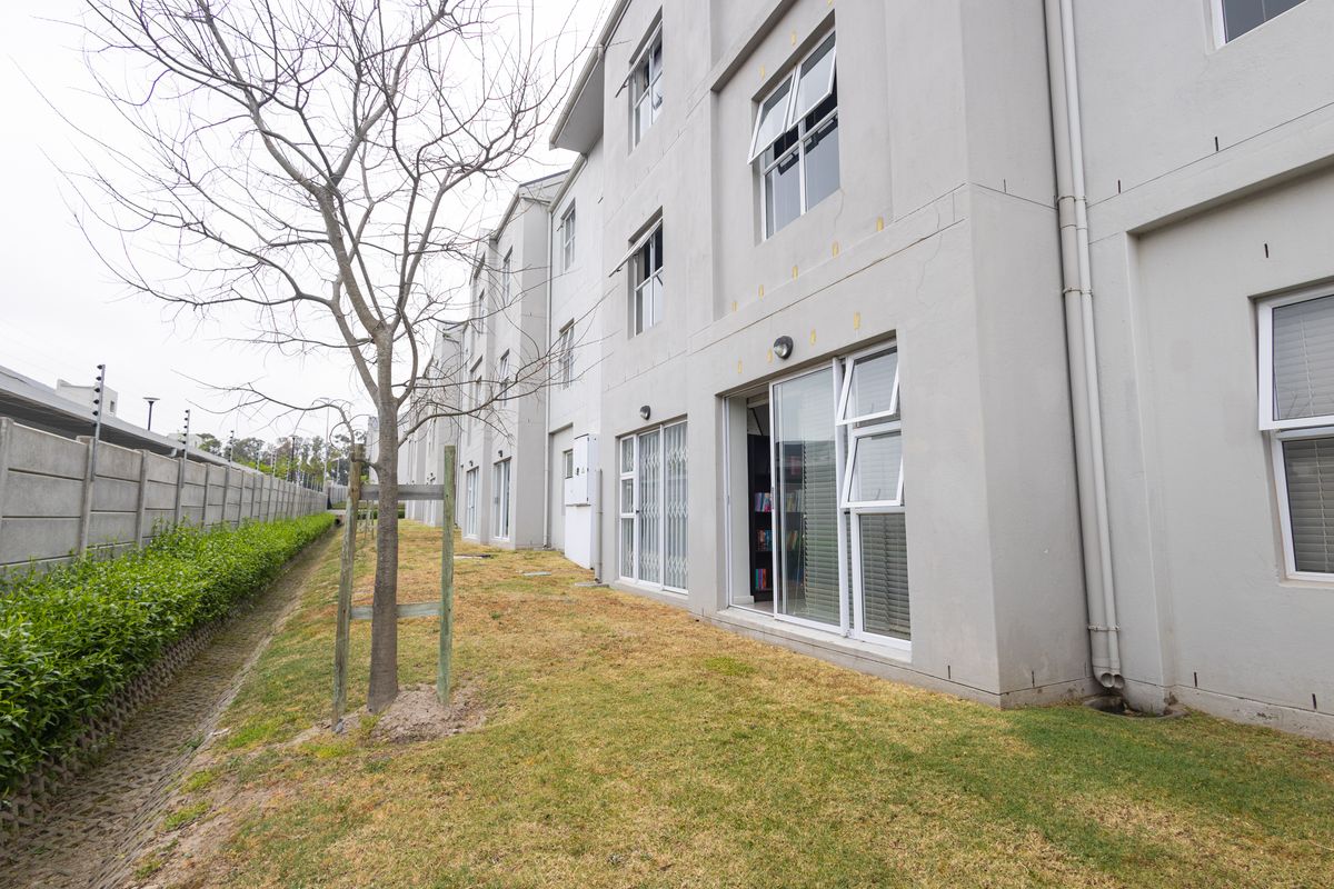 Rear exterior view of a multi-story apartment building featuring sliding glass doors, casement windows, and a landscaped lawn area with boundary wall.
