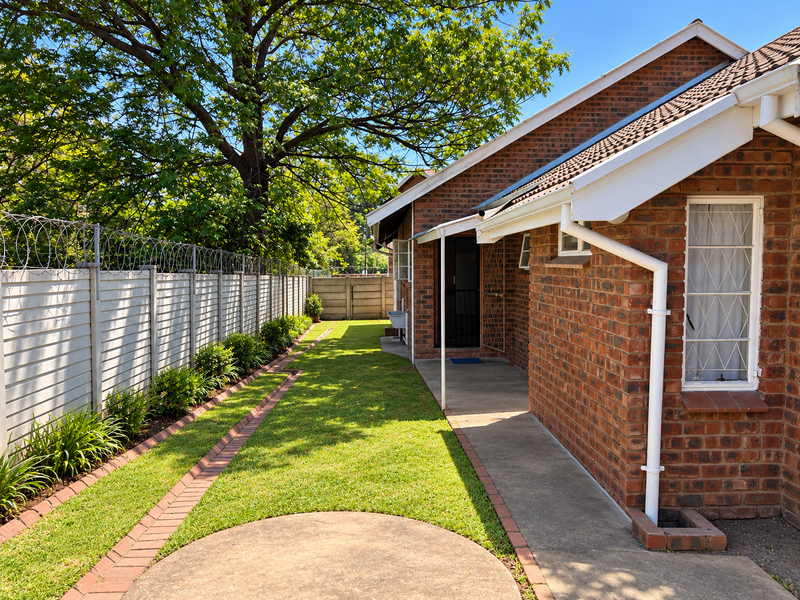 A well-maintained rear exterior view featuring a brick facade, concrete walkway, landscaped garden, and secure boundary wall with razor wire.