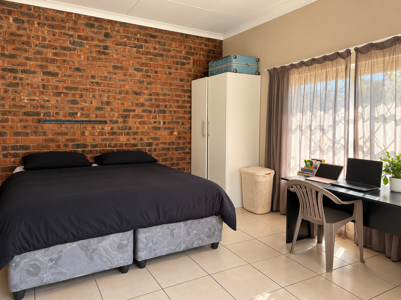 A well-lit bedroom featuring a prominent exposed brick feature wall, neutral tiled flooring, and a large window allowing for natural light.