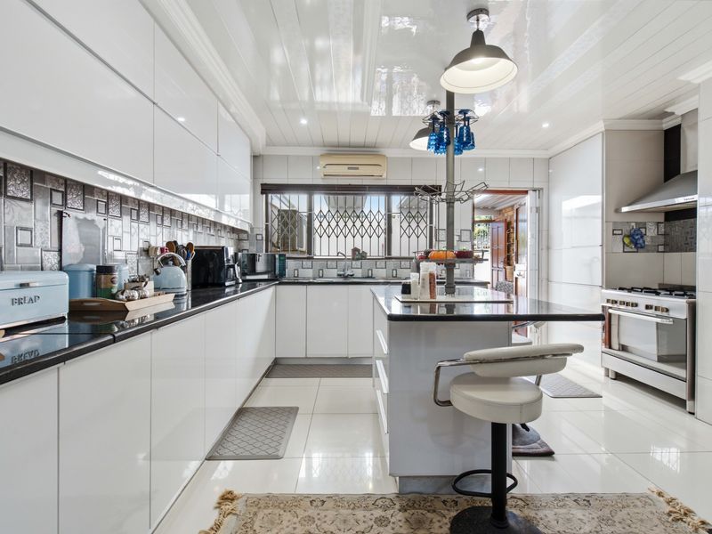 Modern kitchen featuring high-gloss white cabinetry, black granite countertops, a central island, and stainless steel appliances, complemented by polished floor tiles and a glossy ceiling.