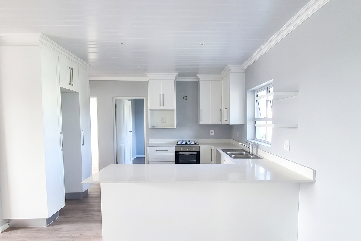 A bright, modern kitchen featuring white cabinetry, stone countertops, integrated oven and hob, and light wood-look laminate flooring in an open-plan layout.