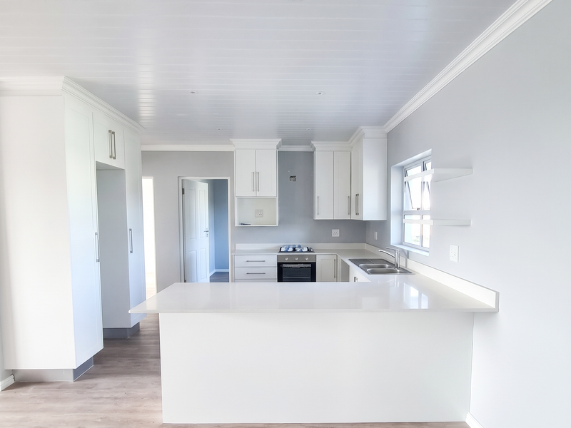 A bright, modern kitchen featuring white cabinetry, stone countertops, integrated oven and hob, and light wood-look laminate flooring in an open-plan layout.