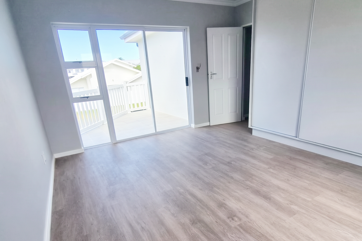 A bright, empty bedroom featuring modern laminate flooring, built-in sliding wardrobes, and direct access to a private balcony through sliding glass doors.