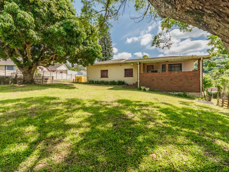 A spacious rear garden featuring a large lawn, mature trees, and a covered patio area with brick detailing.